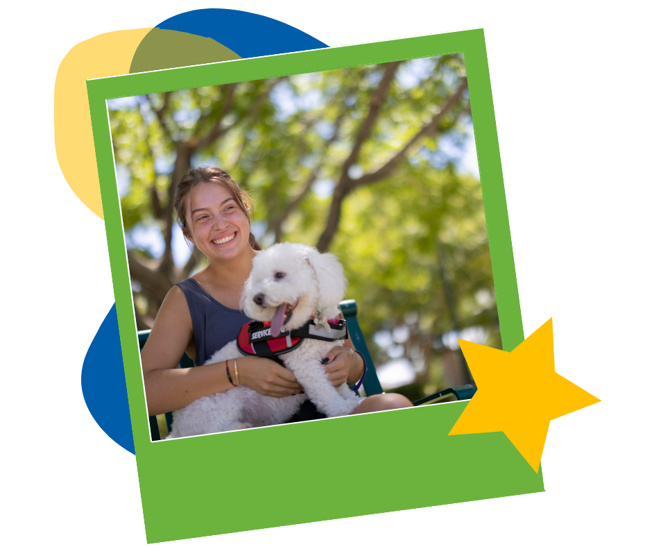 A smiling young woman sitting outdoors under trees with a small white dog in her lap