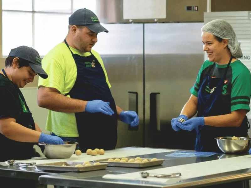 Three bakers in Charley's Cookies aprons and gloves rolling cookie dough at a steel kitchen counter