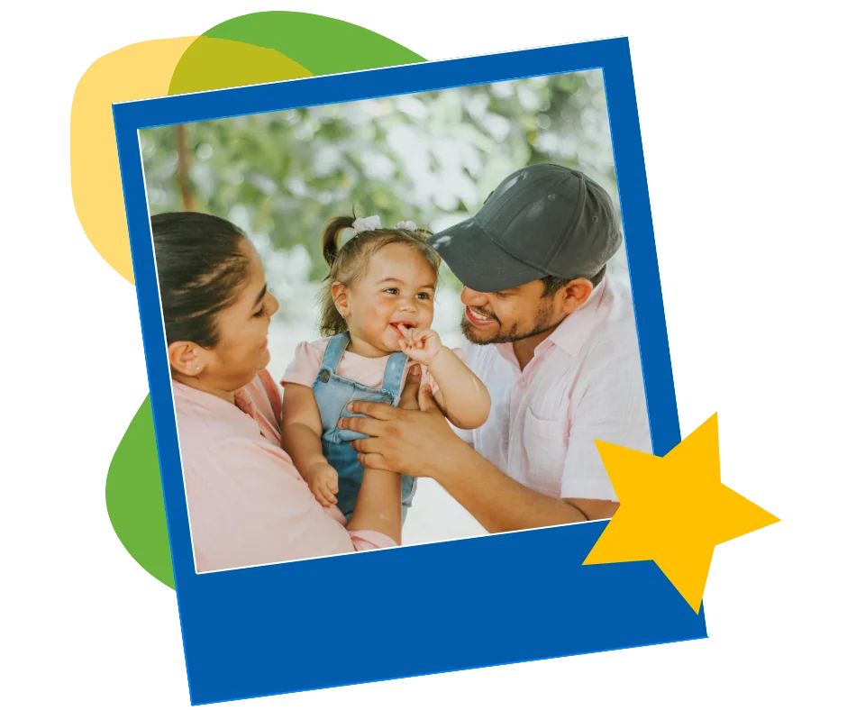 Two parents smiling at a toddler in overalls between them