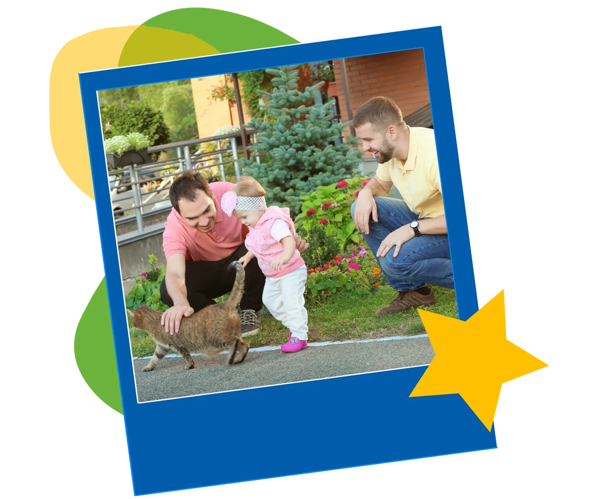 Two men smile as a toddler in a pink headband reaches out to pet a cat on a garden path