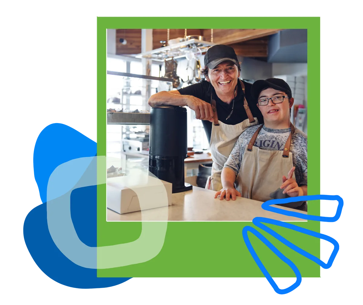 members-cafe-counter-polaroid A smiling man and a young man in aprons lean on a café counter.