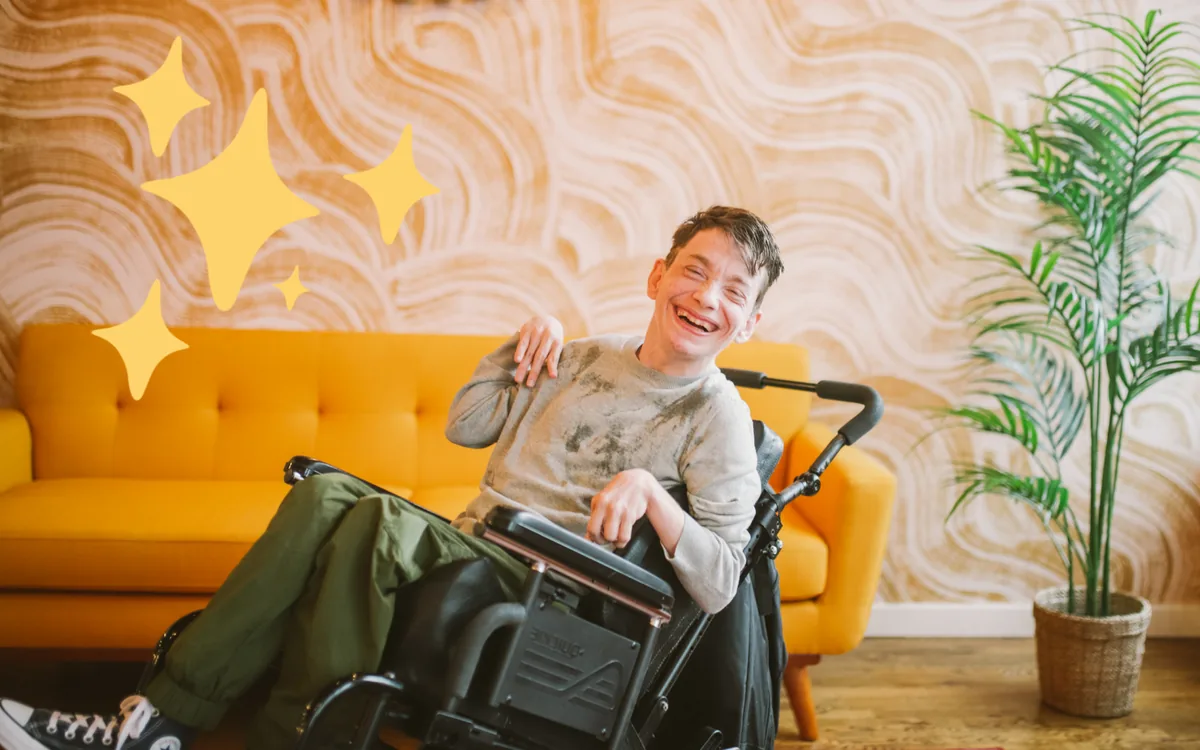 members-laughing-at-home A young man in a wheelchair laughs in a bright living room with an orange sofa.