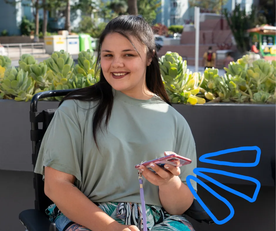 members-smiling-with-phone A young woman in a wheelchair smiles, holding her phone, beside a planter of succulents.