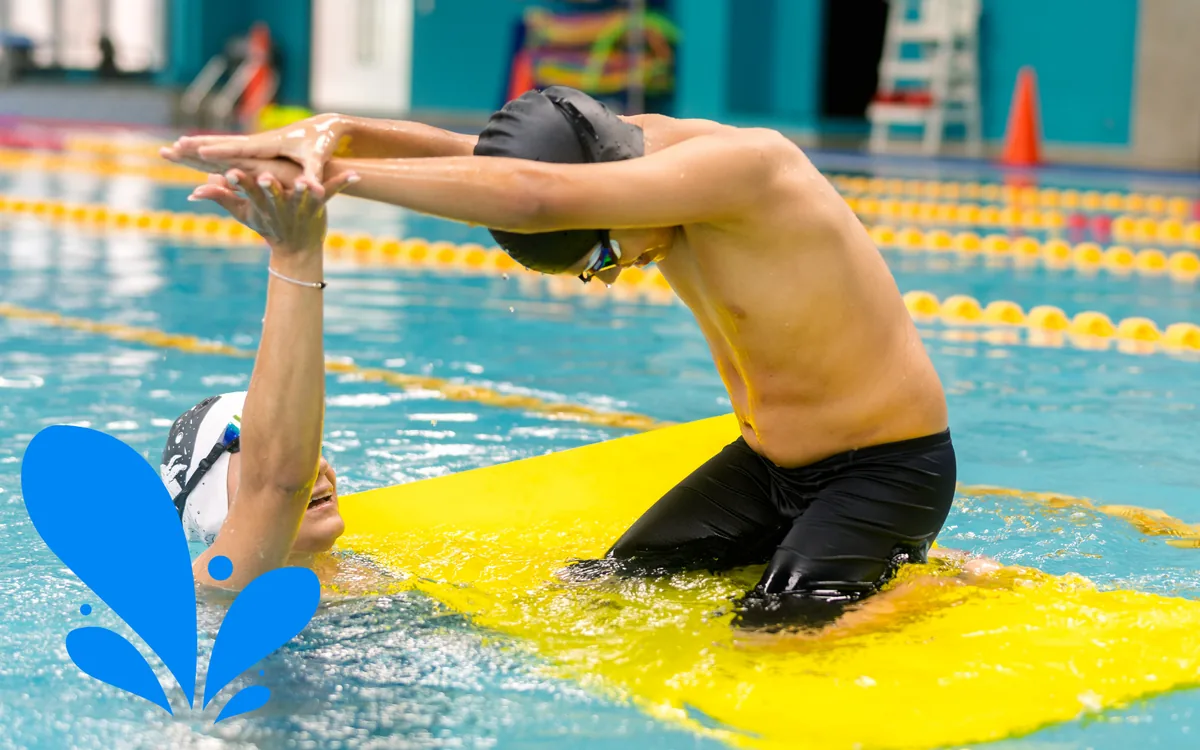 members-swimming-together Two swimmers clasp hands in a pool lane, one kneeling on a yellow mat.