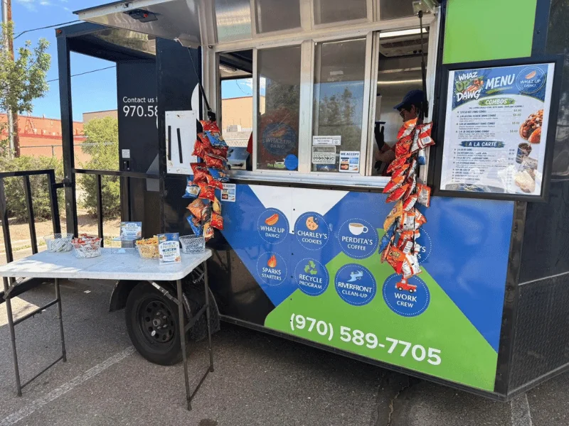 The Whaz Up Dawg food truck with its service window open, a menu board, and snacks hanging by the window